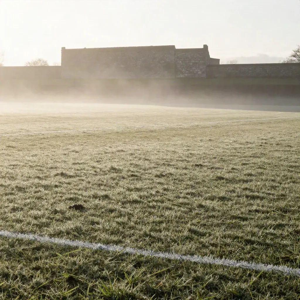 Campo da calcio avvolto nella nebbia all’alba, con linea sbiadita e muretto di pietra sullo sfondo.
